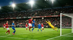 Fodboldbane set fra tribunen med to hold der spiller kamp under projektørlys om aftenen
