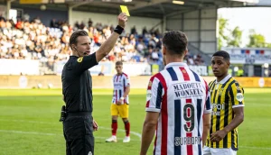 Fodbolddommer der holder et gult kort op under en kamp pa en groenbane med spillere omkring